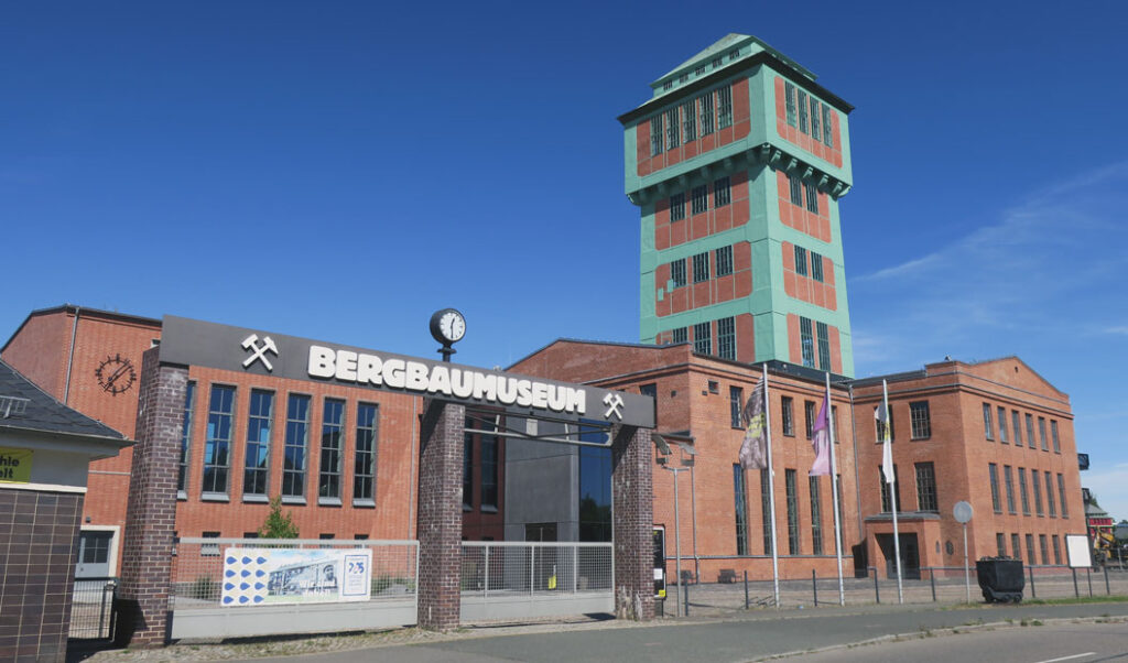 Zu sehen ist der Eingang des Bergbaumuseums KohleWelt. Das Gebäude besteht aus rotem Backstein und hat einen hohen Turm mit einem grünen Dach. Der Turm befindet sich auf der rechten Seite des Gebäudes. Am Eingang befindet sich ein Schild mit der Aufschrift „BERGBAUMUSEUM“ sowie eine Uhr darüber. Auf dem Gelände wehen mehrere Fahnen hinter dem Zaun. Der Himmel ist blau, und im Hintergrund sind einige Wolken zu sehen.