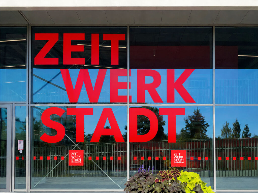 Großflächige Glasfassade mit roter Schrift 'ZEIT WERK STADT' und zwei kleinen roten Schildern mit gleichem Text.