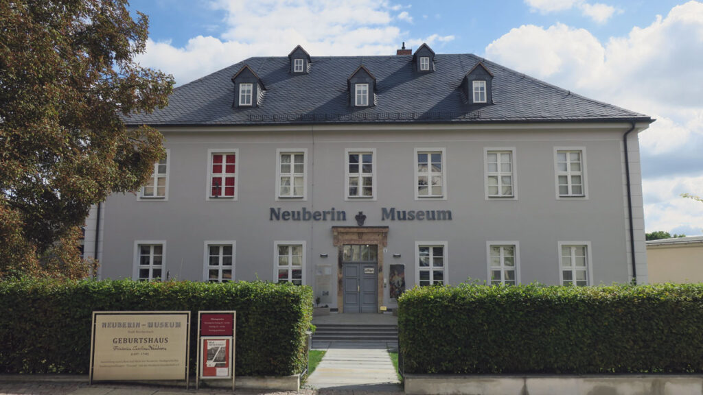 Fassade des Neuberin Museums mit mehreren Fenstern, einem Eingang mit Treppe und einer grünen Hecke im Vordergrund und links zwei Schilder davor.