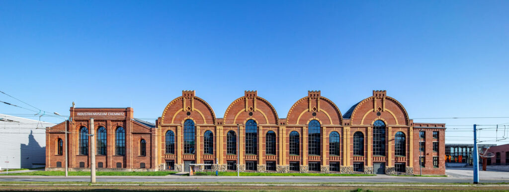 Panoramaansicht eines großen historischen Backsteingebäudes mit mehreren hohen Rundbogenfenstern und markanten Giebeln. Über dem linken Gebäudeteil steht „Industriemuseum Chemnitz“. Im Vordergrund verlaufen Straßenbahngleise, darüber ein klarer blauer Himmel.