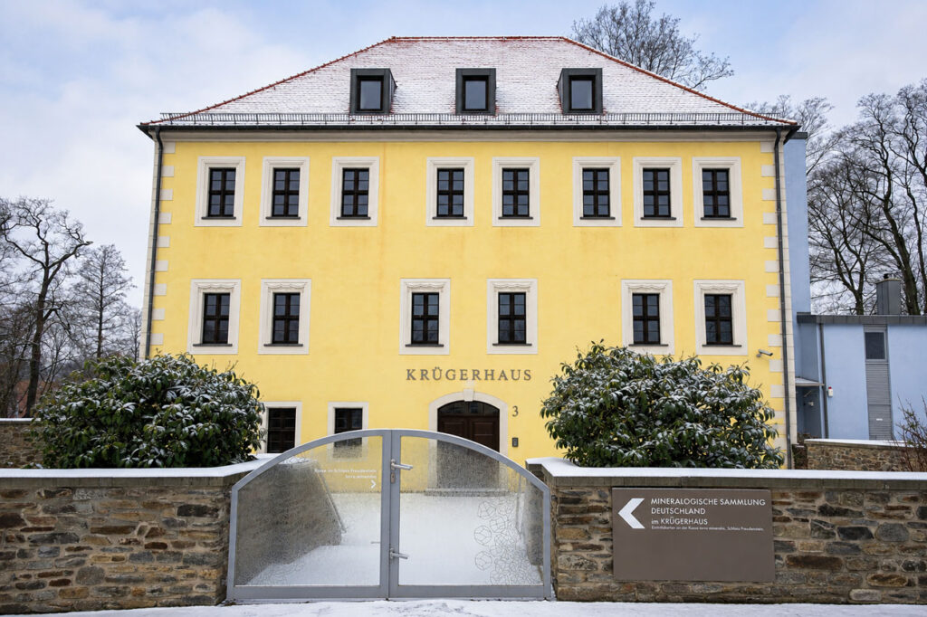 Gelbes dreistöckiges Gebäude mit schwarzem Schriftzug 'KRÜGERHAUS' über dem Eingang und schneebedecktem Dach hinter einer Steinmauer mit Metalltor und zwei Sträuchern.