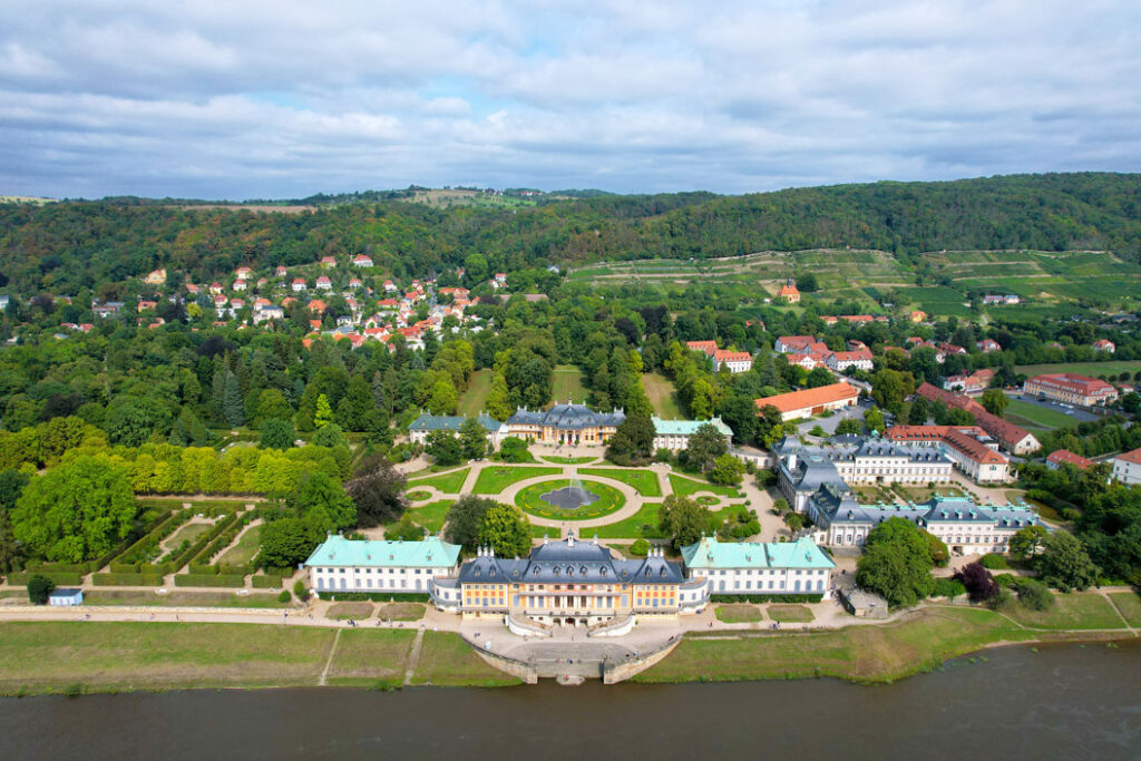 Luftaufnahme des Schlosses Pillnitz mit symmetrischem Garten, umgeben von Wald und der Elbe im Vordergrund.