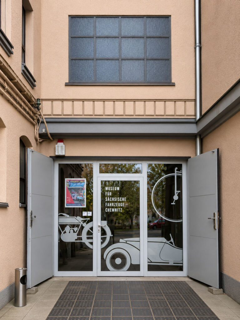 Eingangstür mit geöffneten grauen Flügeltüren und Glasfront mit weißer Grafik eines Oldtimers und Schriftzug 'Museum für sächsische Fahrzeuge Chemnitz'