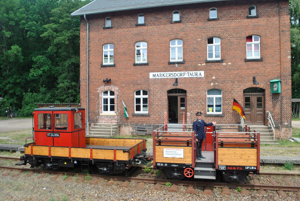 Zwei kleine historische Eisenbahnwaggons auf Gleisen vor einem Backsteingebäude mit Schild 'Markersdorf-Taura' und einem Mann in Uniform auf dem vorderen Waggon