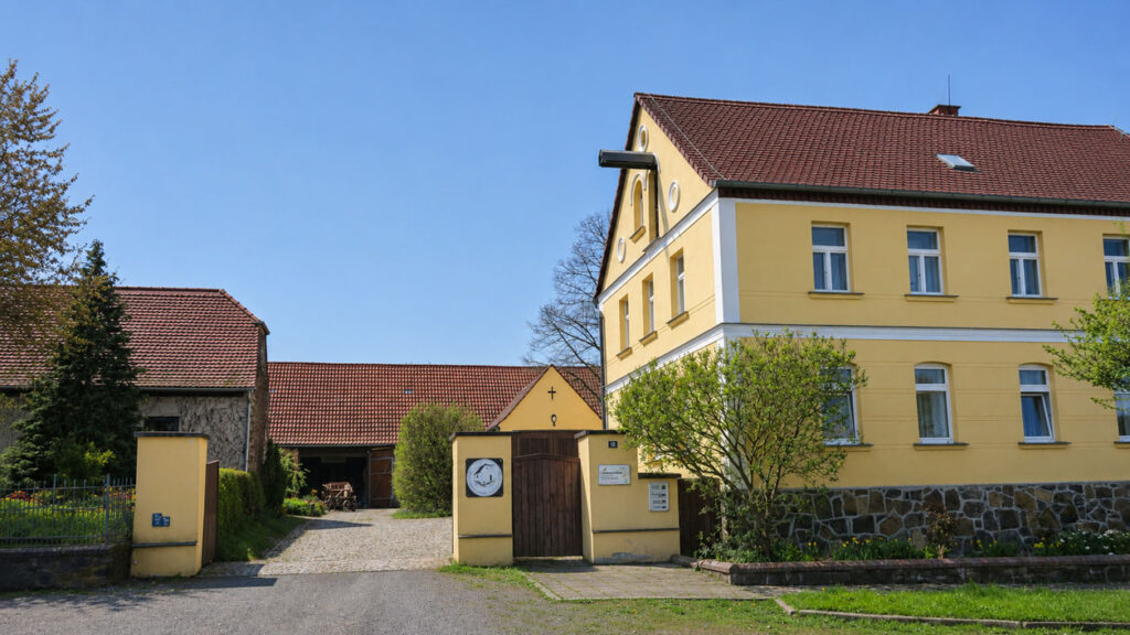 Gelbes zweistöckiges Gebäude mit rotem Ziegeldach neben einem Tor mit Uhr und angrenzendem Hof mit Bäumen