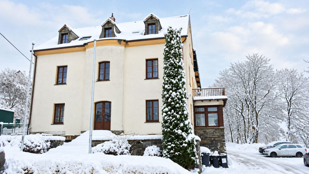 Schneebedecktes mehrstöckiges Gebäude mit Satteldach, mehreren Fenstern und einem hohen Nadelbaum davor