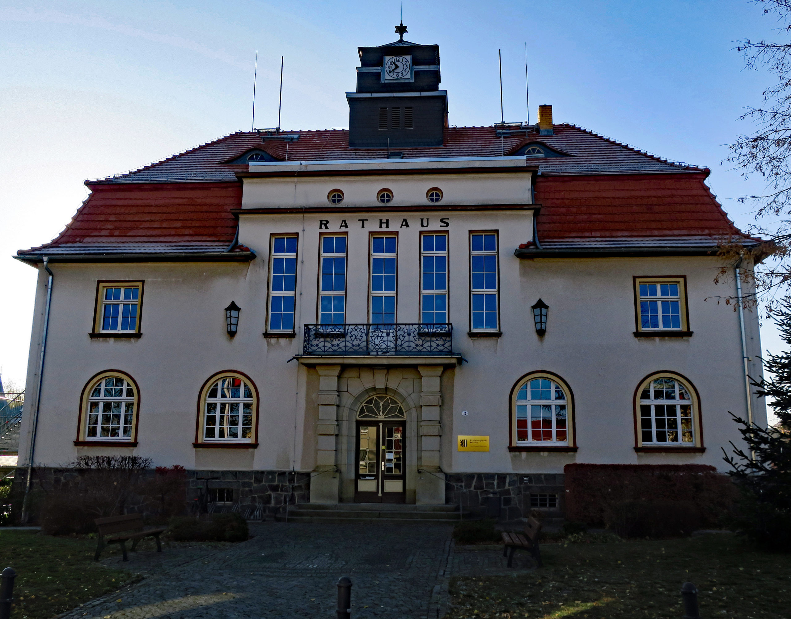 Das Rathaus Weixdorf mit Turm und Uhr nimmt den größten Teil des Bildes ein. Der Sockel des Gebäudes besteht aus Sandstein und hat zwei sichtbare Kellerfenster. In der Mitte befindet sich die Eingangstür mit Rundbogen und davor drei Stufen, welche in das Rathaus führen. Die erste Etage hat rechts und links neben der Tür jeweils zwei Bogenfenster. In der zweiten Etage gibt es mittig einen Balkon, auf diesen gelangt man durch eine sehr schmale hohe Balkontür. Links und rechts davon gibt es jeweils zwei sehr schmale hohe rechteckige Fenster, eine Laterne und ein quadratisches Fenster auf jeder Seite. Vom unteren Bildrand führt mittig ein gepflasterter Weg zum Rathaus. Links und rechts davon ist Rasenfläche und auf beiden Seiten jeweils eine Holzbank am Rand des Weges. Unmittelbar vor dem Rathaus sind auf der rechten und linken Seite Hecken gepflanzt.