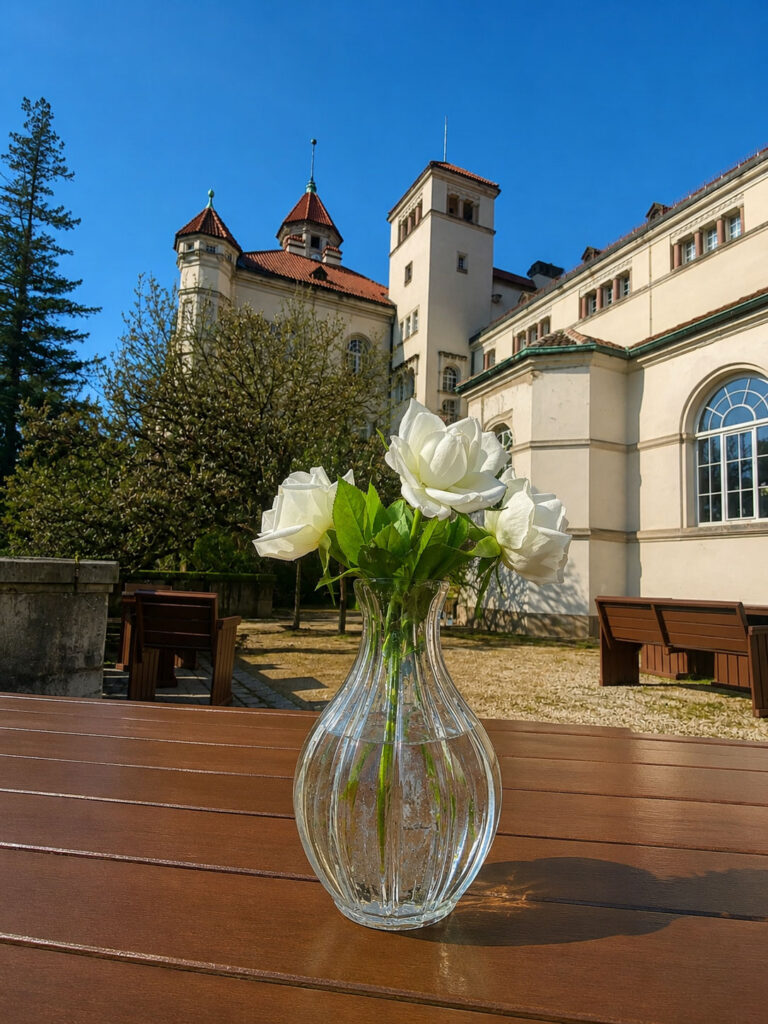 Vase mit weißen Rosen auf Holztisch vor Schloss mit Türmen und Bäumen im Hintergrund