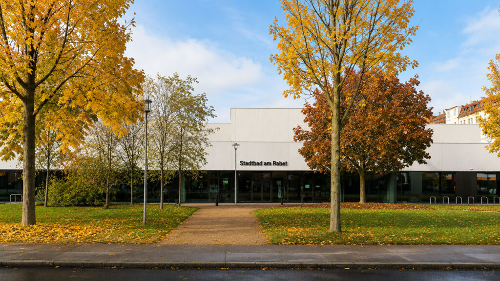 Außenansicht des Stadtbads am Rabet in Leipzig. Das moderne Gebäude ist von herbstlich gefärbten Bäumen umgeben, ein gerader Weg führt zum Haupteingang.