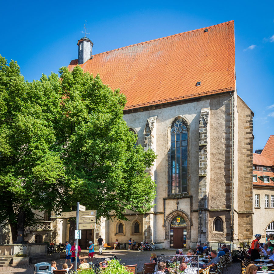 Historisches Gebäude mit großem spitzbogigem Fenster und rotem Satteldach neben einem Platz mit Bäumen und Menschen