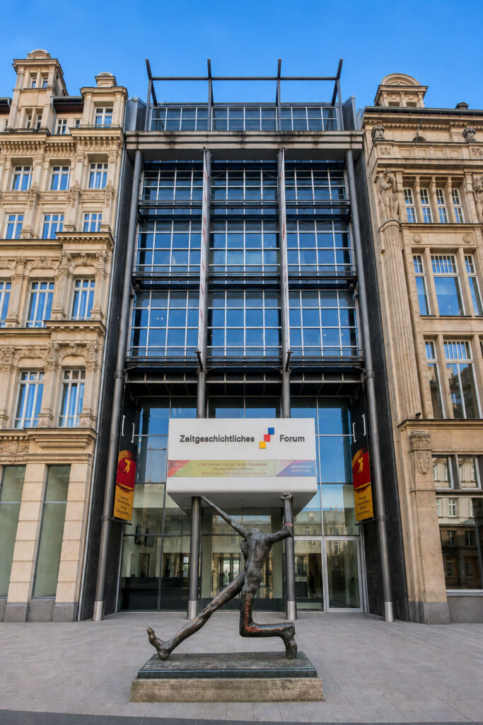 Fassade eines historischen Gebäudes mit moderner Glasfront und einer bronzenen Skulptur einer knienden Figur mit erhobenen Armen davor